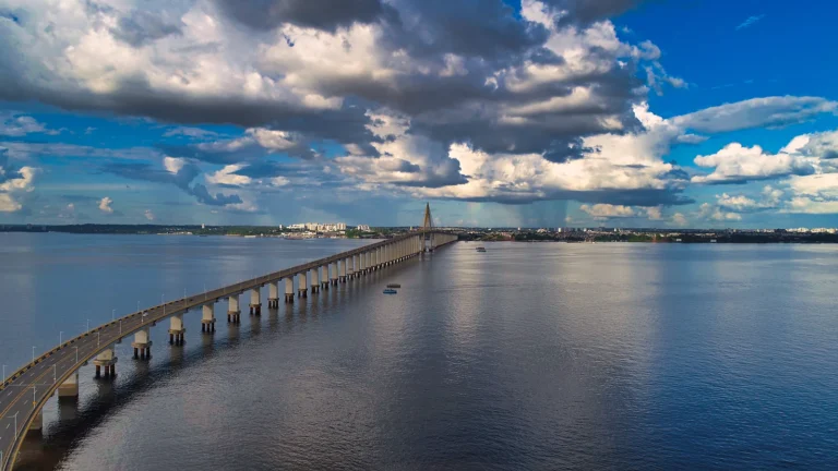 Ponte Rio Negro atravessando o rio em diagonal, com pilares refletidos na água escura e nuvens grandes num céu azul sobre Manaus ao fundo