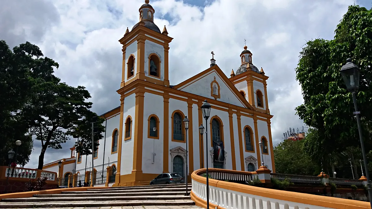 Fachada branca com detalhes amarelos da Catedral Metropolitana de Manaus, a Igreja Matriz, com duas torres, escadaria e árvores dos dois lados, sob céu com nuvens