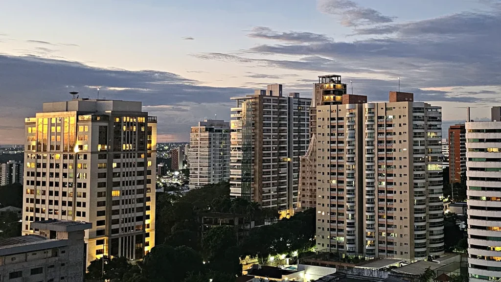 Prédios do bairro Adrianópolis, em Manaus, ao entardecer, com a fachada de uma torre iluminada pelo sol e janelas acesas