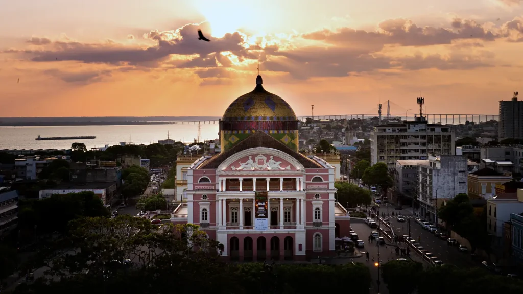 Teatro Amazonas visto do alto ao pôr do sol, com a fachada rosa, a cúpula colorida e o rio Negro com a ponte ao fundo