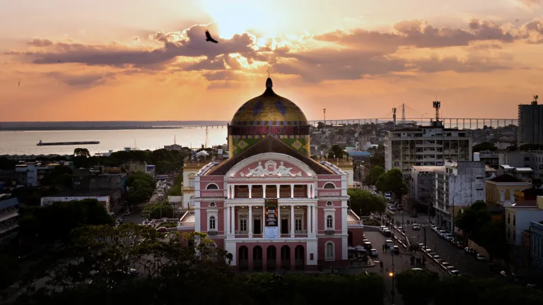 Teatro Amazonas visto do alto ao pôr do sol, com a fachada rosa, a cúpula colorida e o rio Negro com a ponte ao fundo