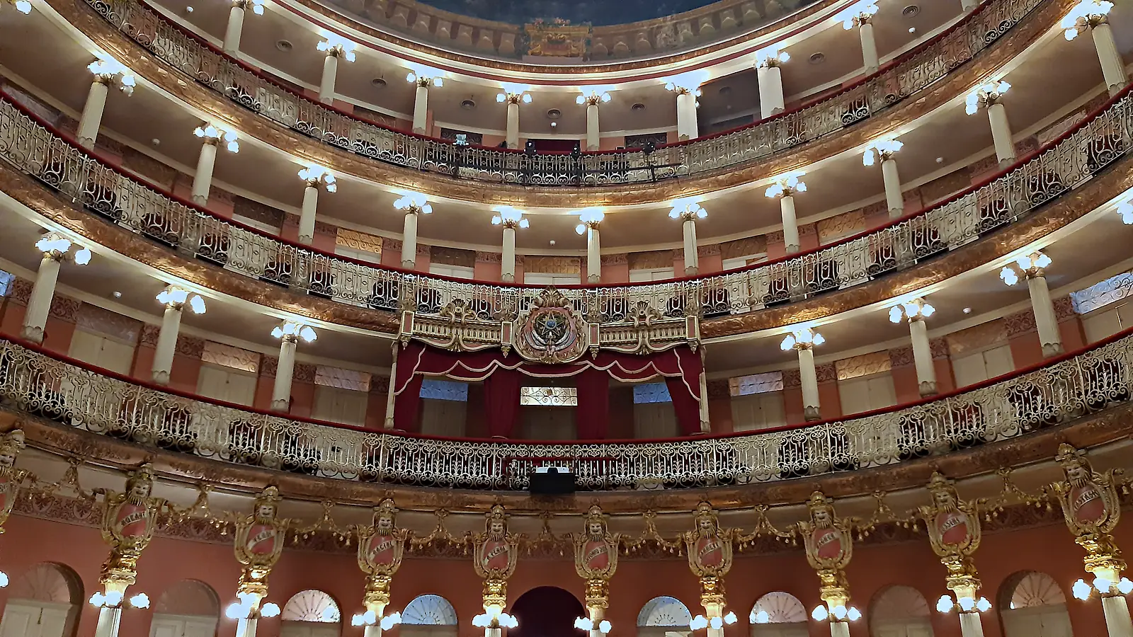 Sala de espetáculos do Teatro Amazonas vista da plateia, com três andares de galerias curvas de ferro dourado, colunas iluminadas e o camarote central com cortina vermelha
