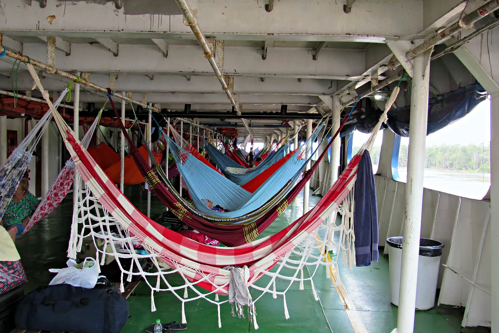 Redes coloridas armadas lado a lado no convés coberto de um barco de recreio que navega no rio Amazonas, com bagagens no chão