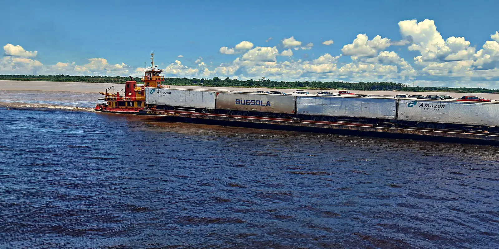Balsa de carga empurrada por rebocador no rio Amazonas perto de Manaus, levando carretas e carros enfileirados no convés, em dia de sol