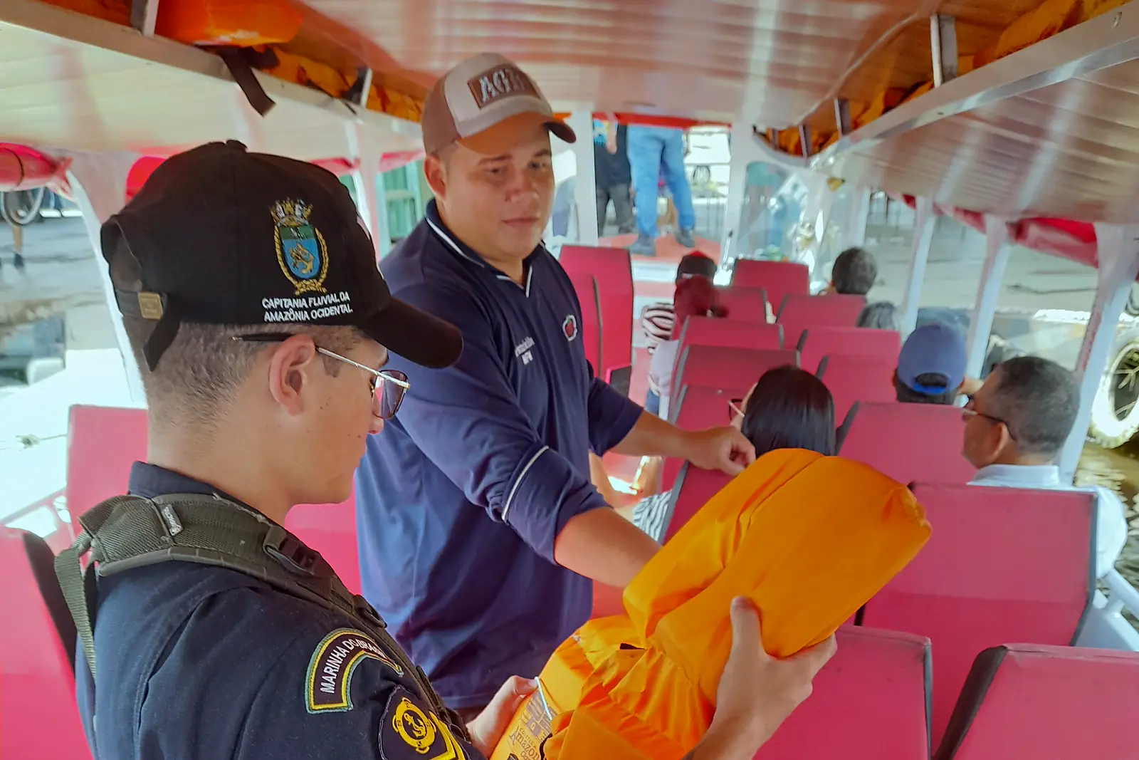 Cabine de passageiros com fileiras de poltronas vermelhas e coletes salva-vidas laranja guardados no teto, durante a inspeção de um militar da Capitania Fluvial da Amazônia Ocidental em Manaus