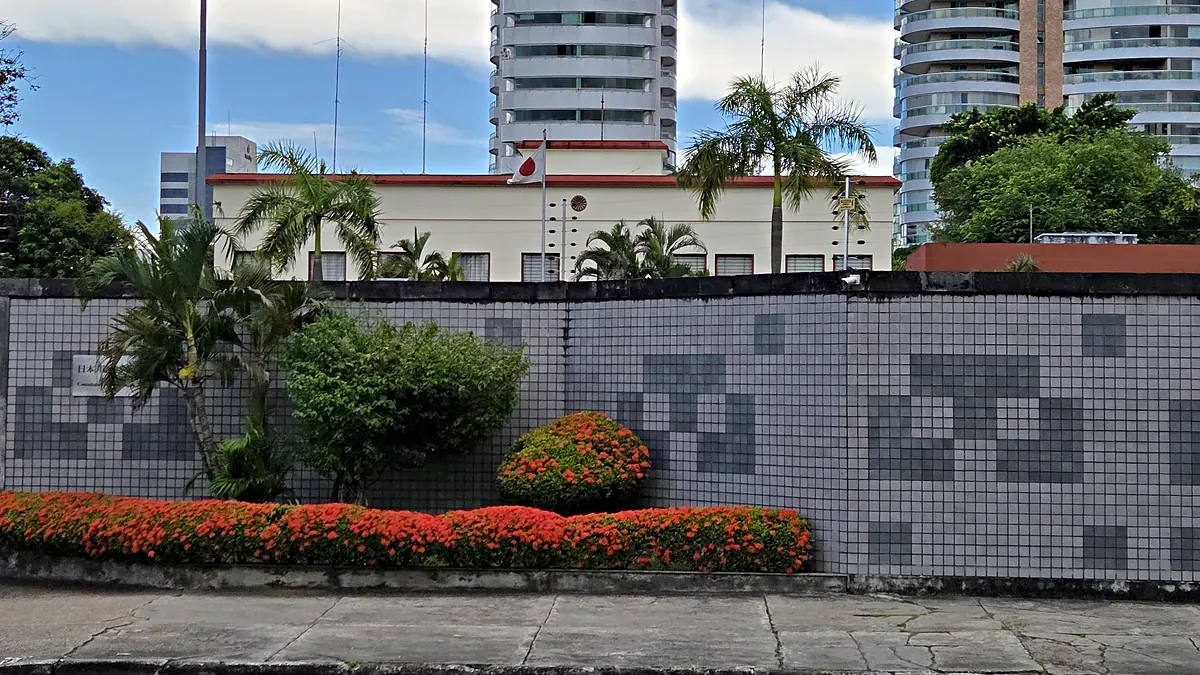 Prédio branco do Consulado-Geral do Japão em Manaus com a bandeira do Japão no mastro, atrás de um muro de pastilhas cinza com canteiro de flores vermelhas, em Adrianópolis
